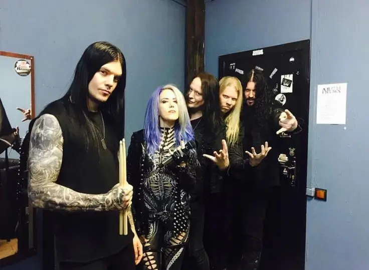 Arch Enemy band members backstage, with Alissa White-Gluz in front, drummer holding sticks, and all members dressed in black posing for the camera