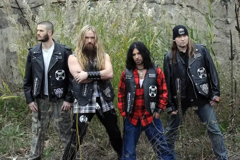 Black Label Society posing outdoors in leather vests and flannel, standing against a rugged backdrop of overgrown grass and weathered walls.