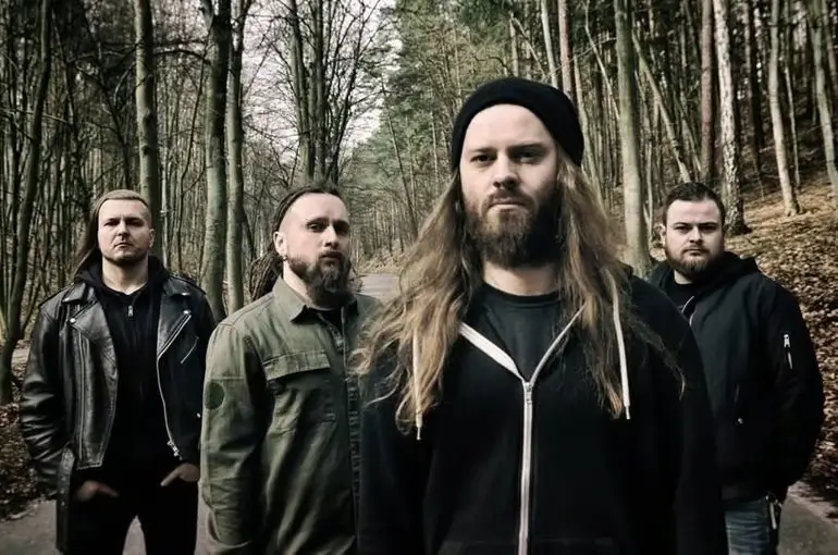 Decapitated posing in a forest, dressed in dark clothing, with a serious and intense expression.