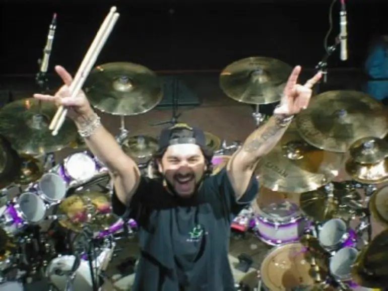 Mike Portnoy raises his drumsticks and throws devil horns, surrounded by his drum kit.