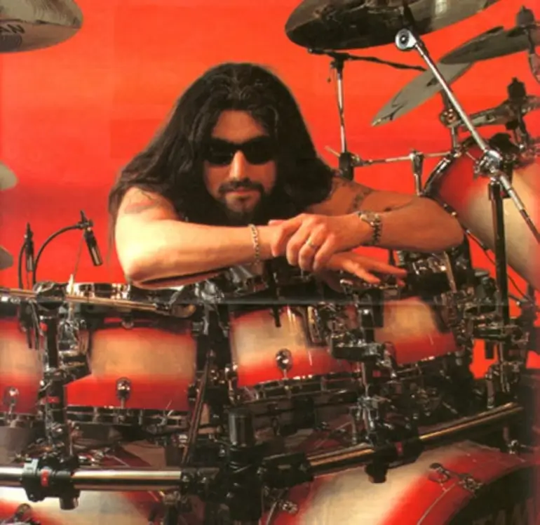 Mike Portnoy in sunglasses, leaning on a red drum set with a serious expression.