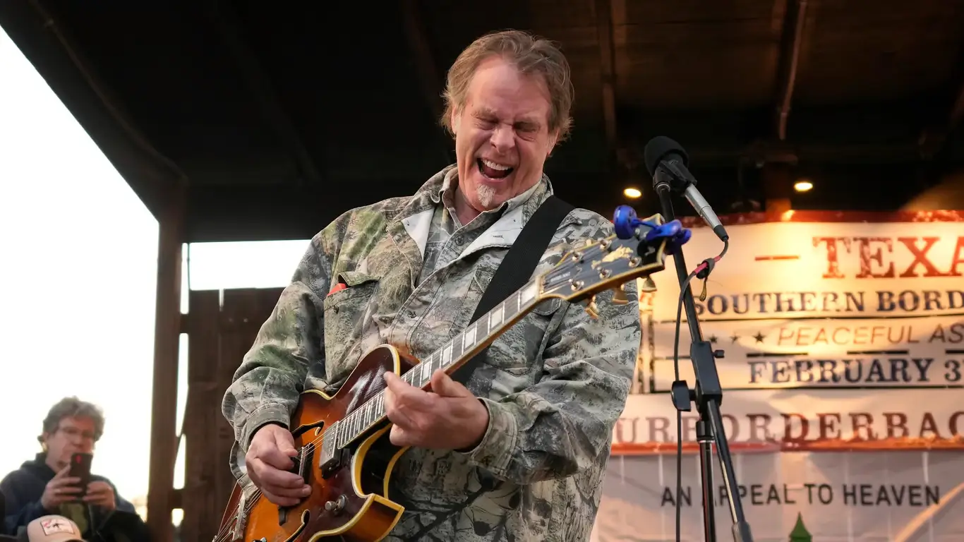 Ted Nugent performing live on stage, wearing camouflage clothing and playing a Gibson Byrdland guitar.